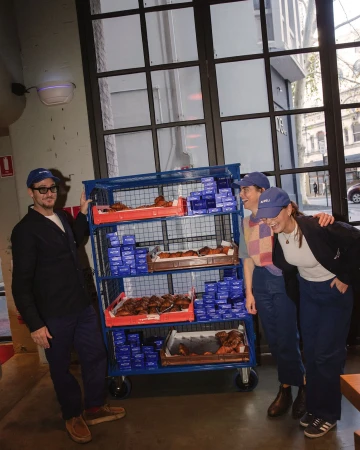 Group of three adults in matching hats standing with a metal cart filled with croissants and blue boxes in an indoor urban cafe setting.