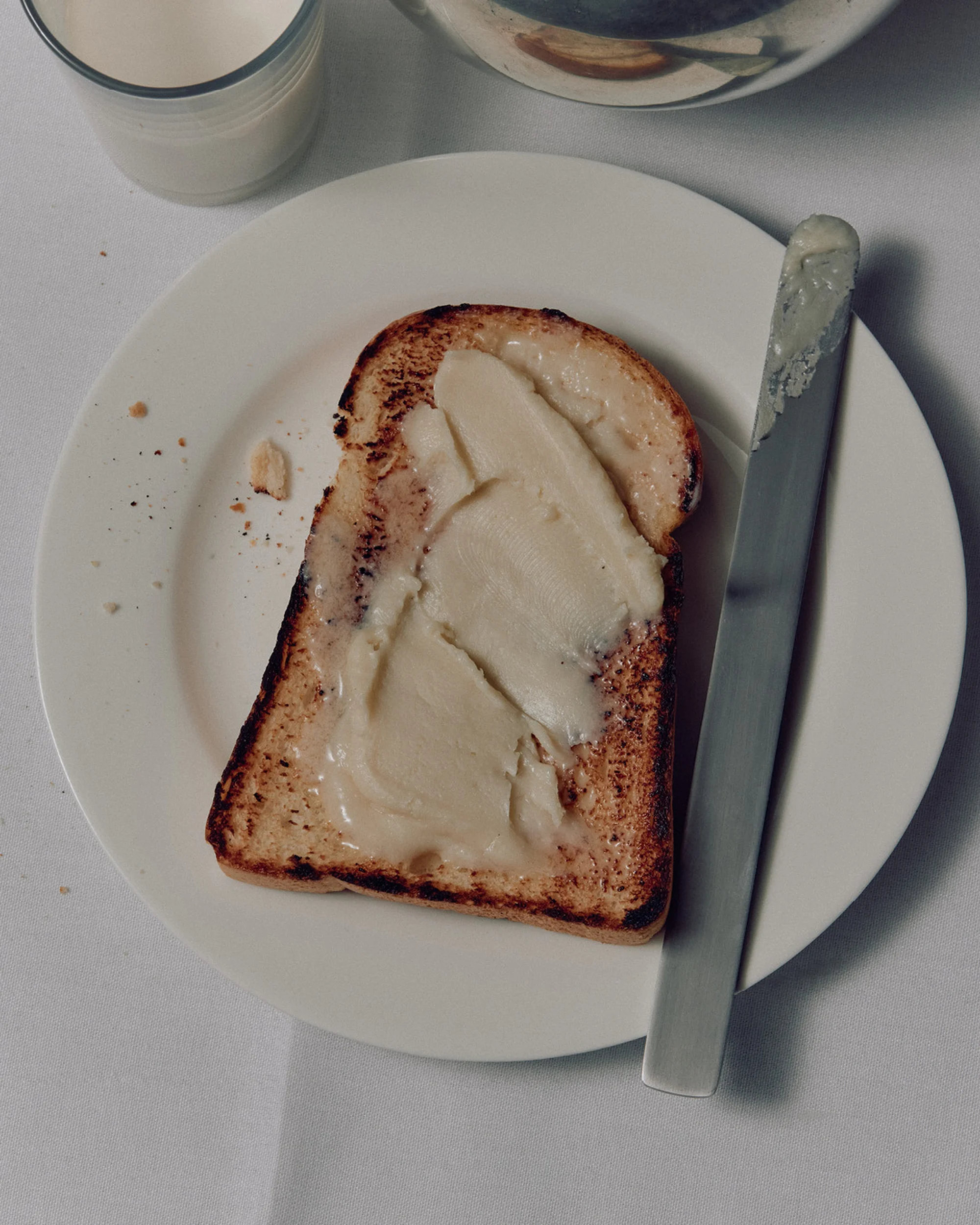 Overhead shot of toasted bread with butter on a white plate, alongside a knife and glass of milk on a white surface.