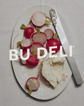 Overhead view of fresh radishes and white cheese on an oval white plate with a knife, featuring the Bu Deli brand logo.