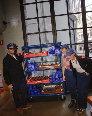 Three people in matching blue caps posing beside a wheeled bakery cart filled with pastries and blue boxes