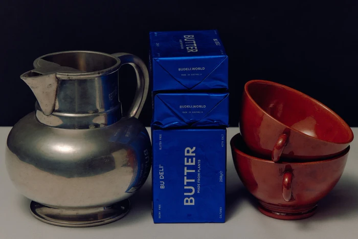 A silver metal pitcher, three stacked blue butter packages, and two stacked red ceramic cups on a white surface.