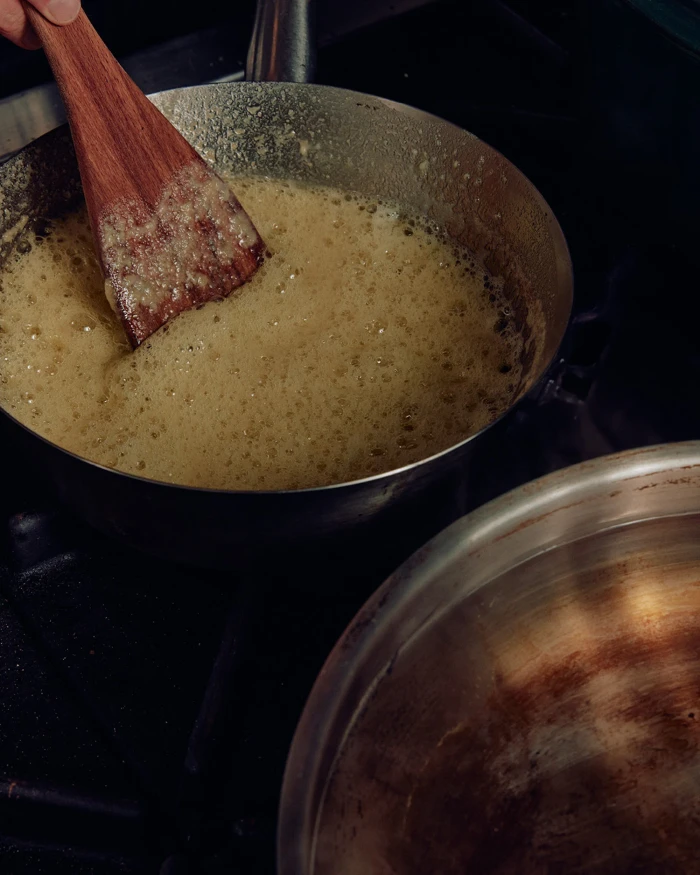 Close-up of a wooden spatula stirring bubbling golden liquid in a metal saucepan on a dark stovetop