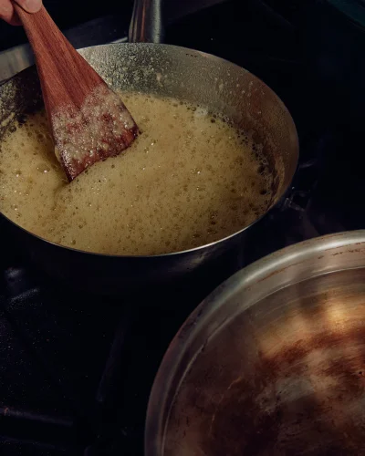Close-up of a wooden spatula stirring bubbling golden liquid in a metal saucepan on a dark stovetop