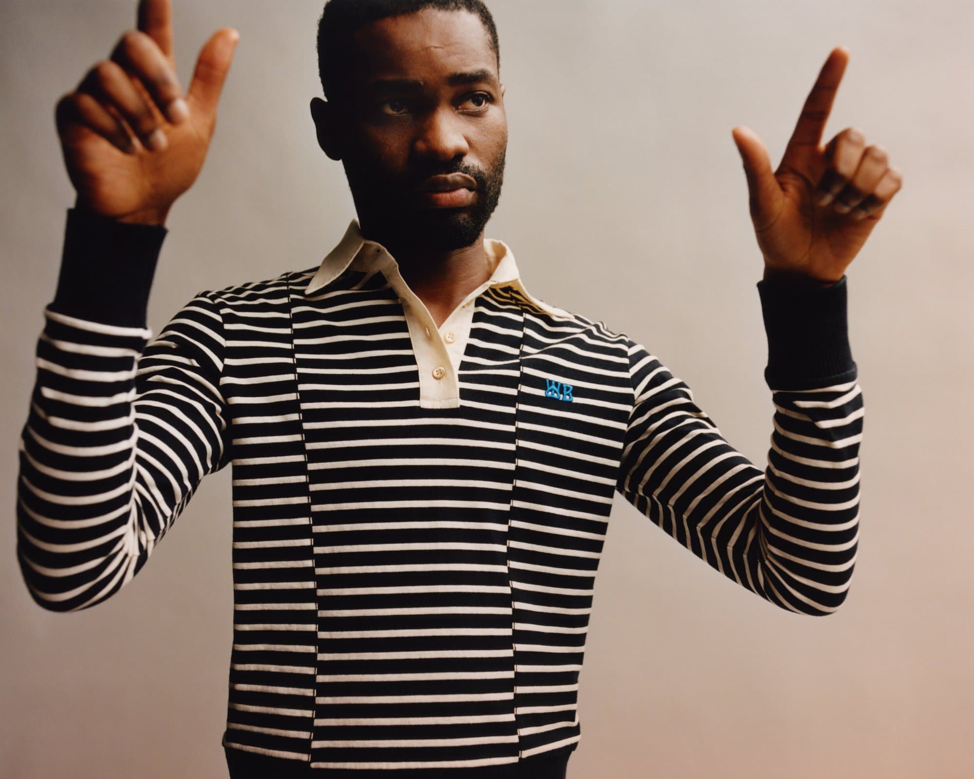 A man with a serious expression in a black and white striped long-sleeve shirt raises his hands in an indoor setting with neutral lighting.
