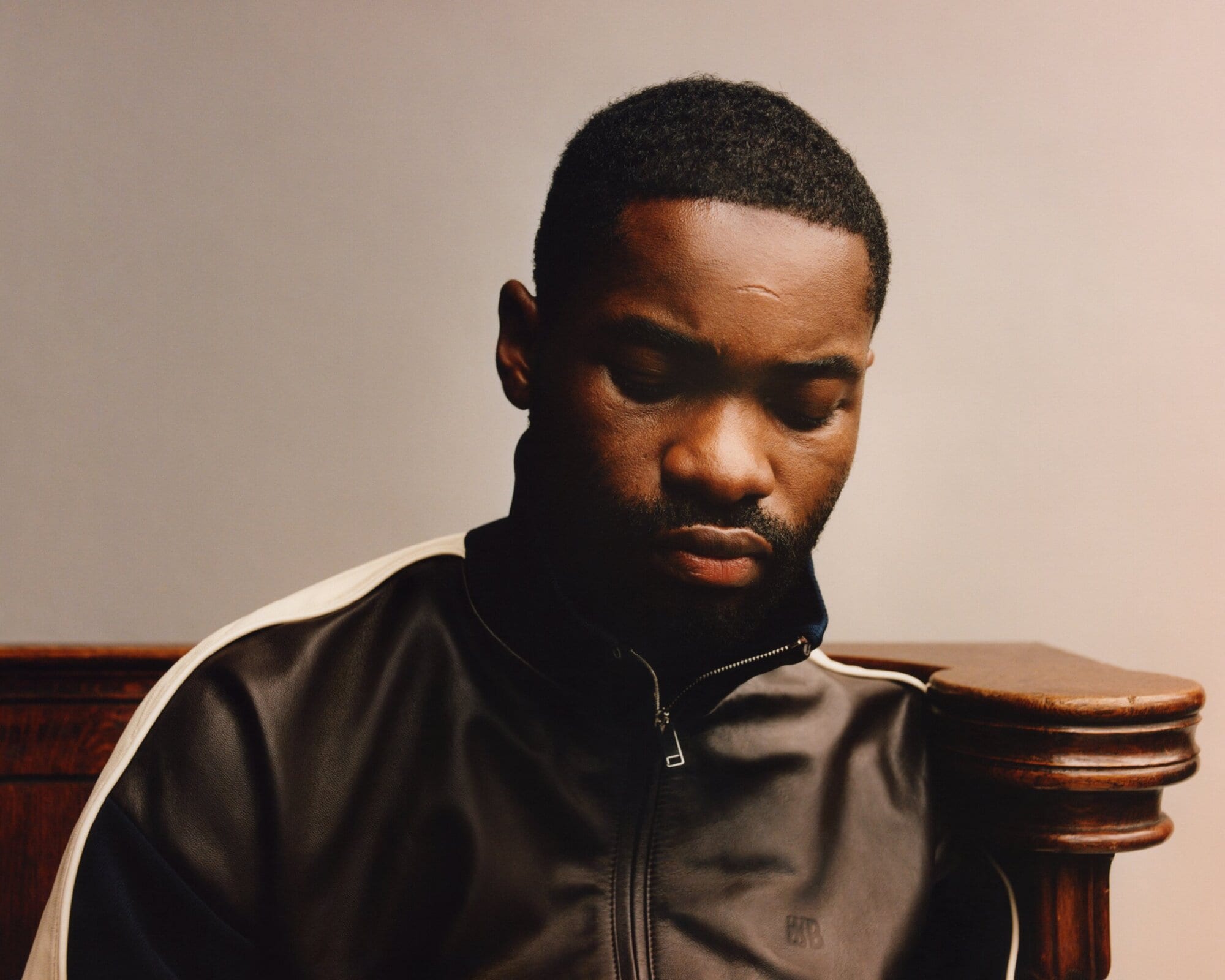 Portrait of an adult Black man looking down, wearing a black and white jacket, captured in a studio setting with neutral lighting.