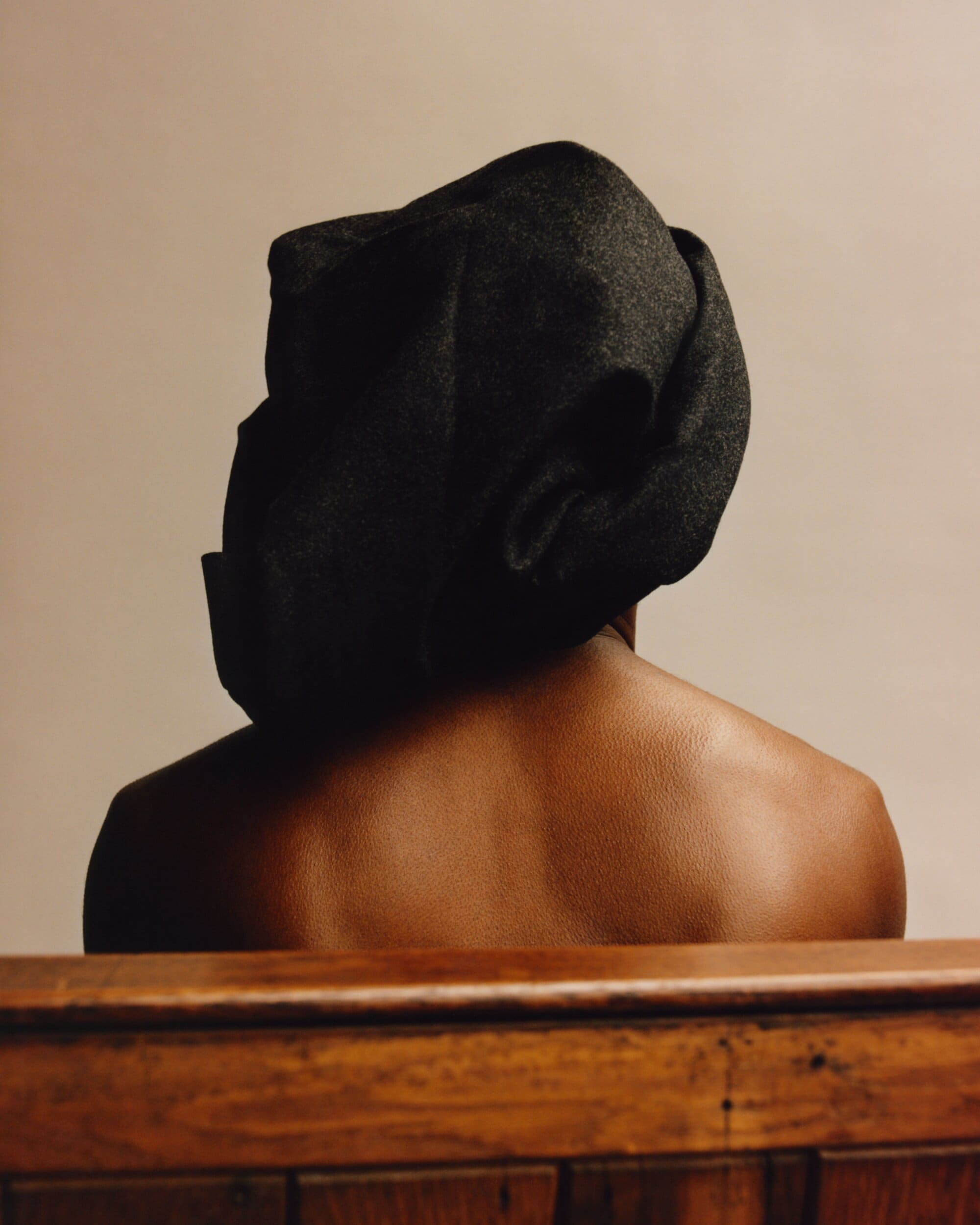 Back view of an African American adult wearing a black head covering, revealing bare shoulders and wooden paneling in the foreground.