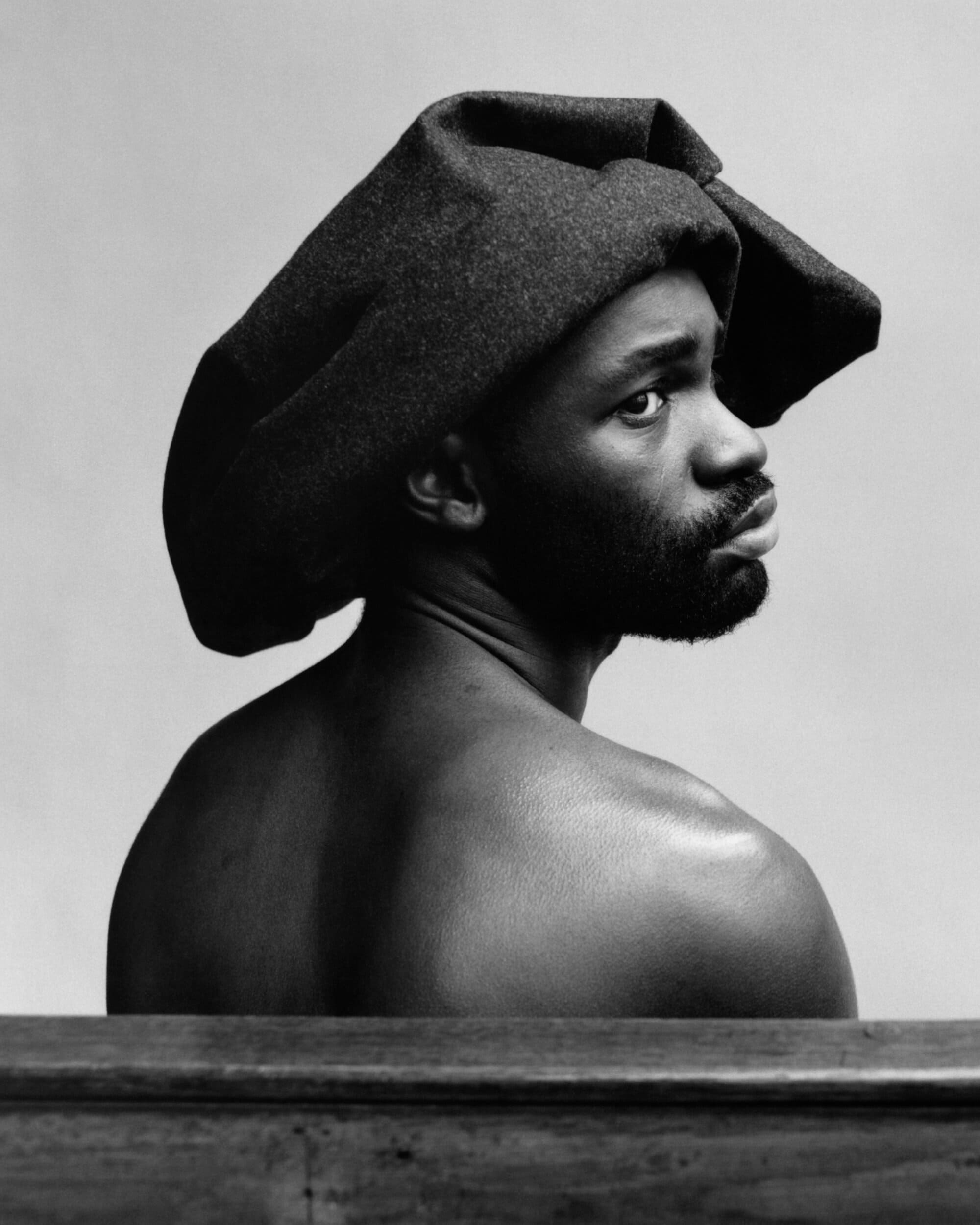 Monochrome portrait of a Black man with dark facial hair wearing a textured dark grey head wrap looking over his shoulder.