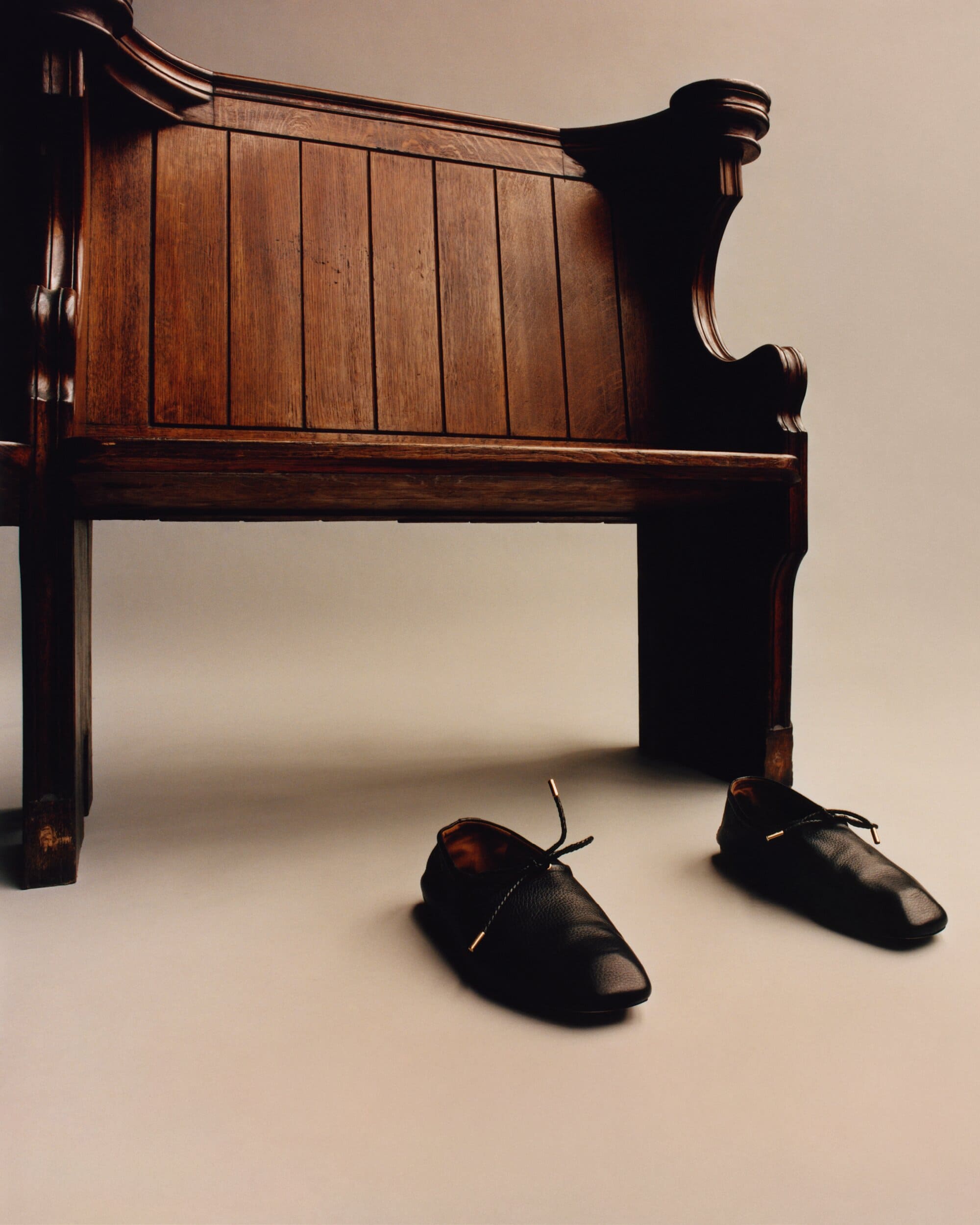 Still life shot of a vintage wooden pew with architectural details and a pair of black leather lace-up shoes on a gray studio backdrop.