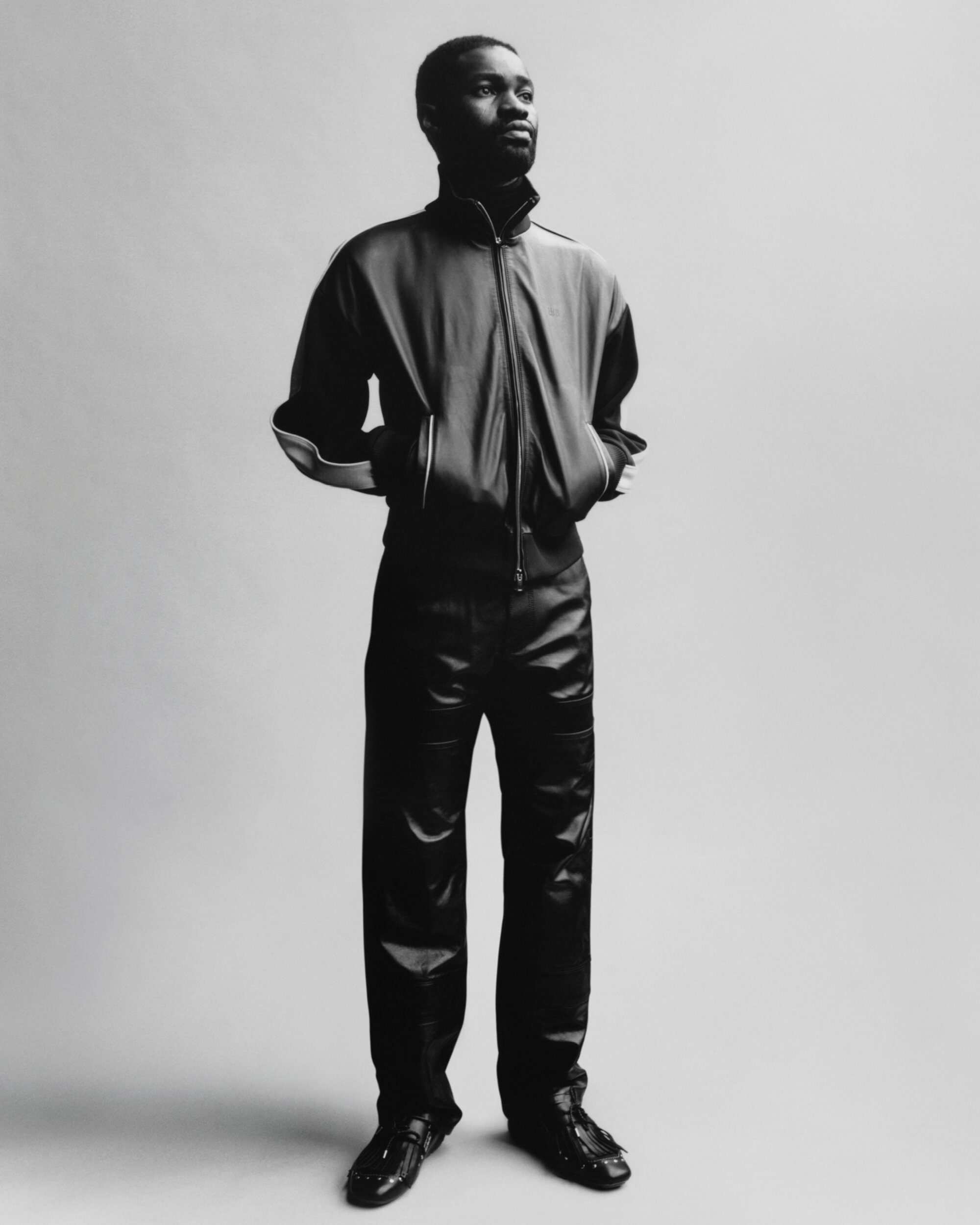 A Black man in a shiny tracksuit stands posing against a plain background in a black and white studio portrait.