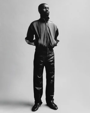 Black and white portrait of a man standing against a plain background wearing a dark zipped jacket and trousers