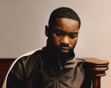 Close-up portrait of a Black man with short hair wearing a dark zip-up track jacket against a neutral studio background