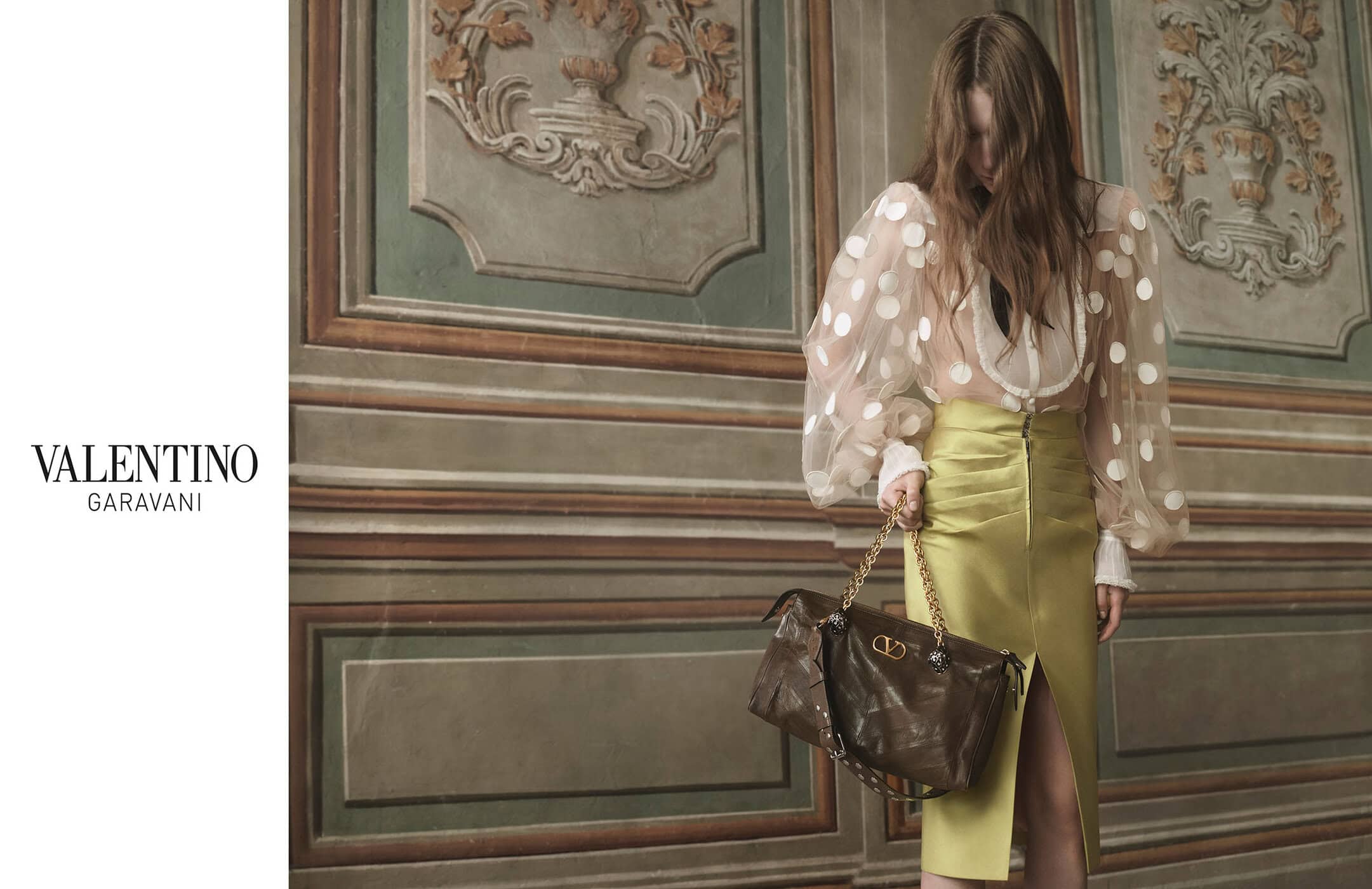 Woman in polka dot blouse and yellow skirt carrying a leather handbag with gold chain, posing in front of a decorative wall.