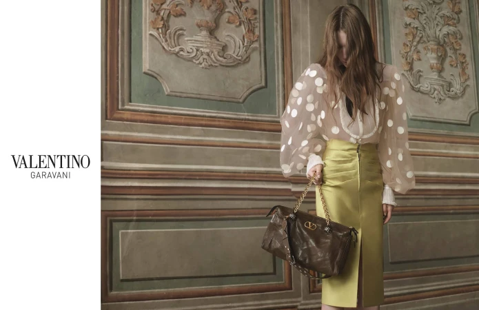 Woman in Valentino Garavani silk skirt and polka dot blouse holding a brown handbag stands in front of an ornate wall.
