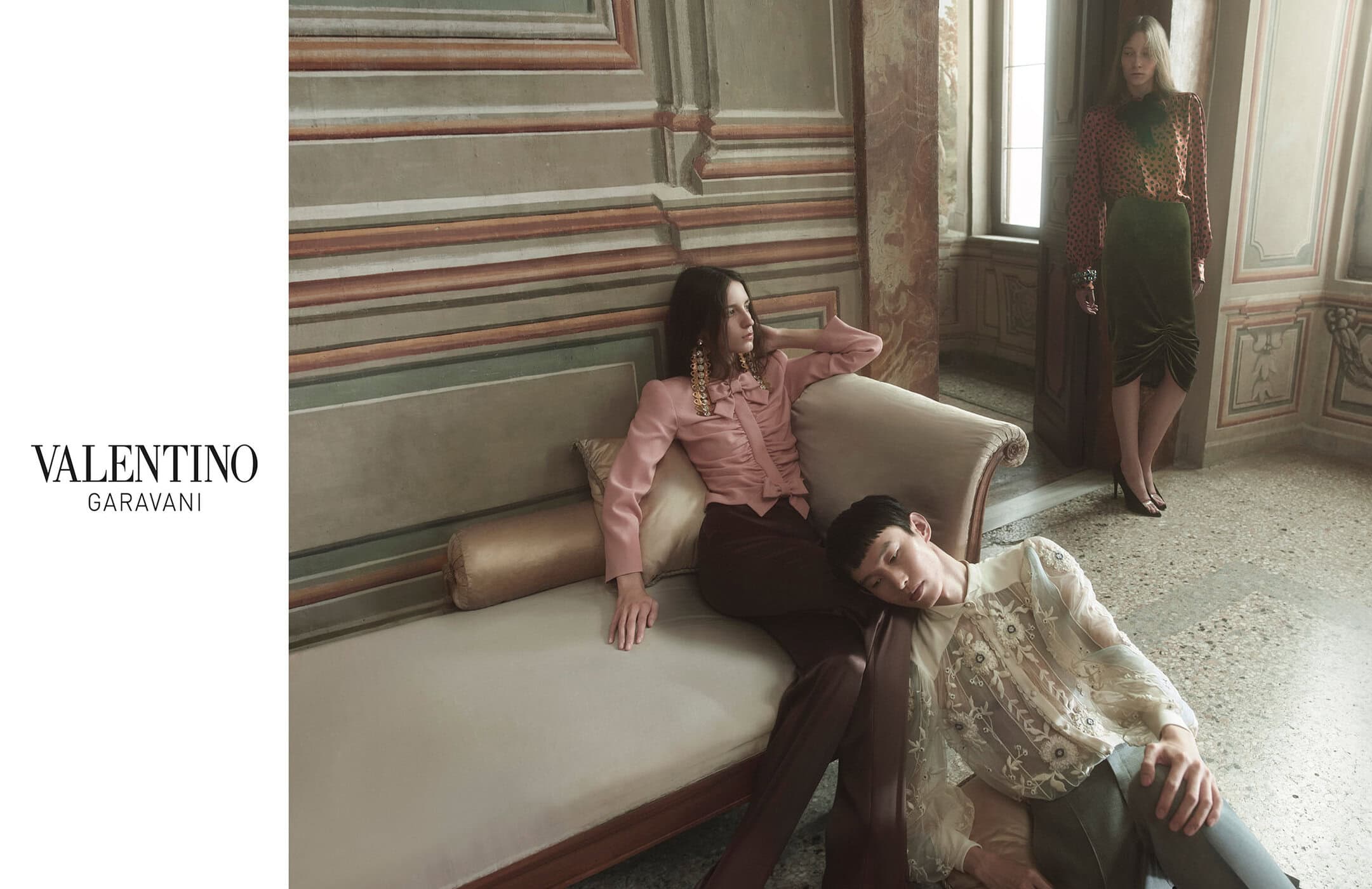 Three fashion models in a room with a wall mural wearing Valentino Garavani clothing; two are on a sofa; editorial fashion shot.
