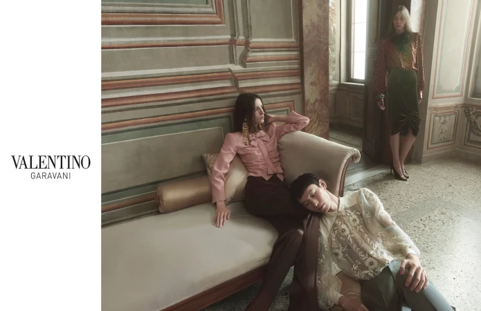 Three fashion models in a room with a wall mural wearing Valentino Garavani clothing; two are on a sofa; editorial fashion shot.