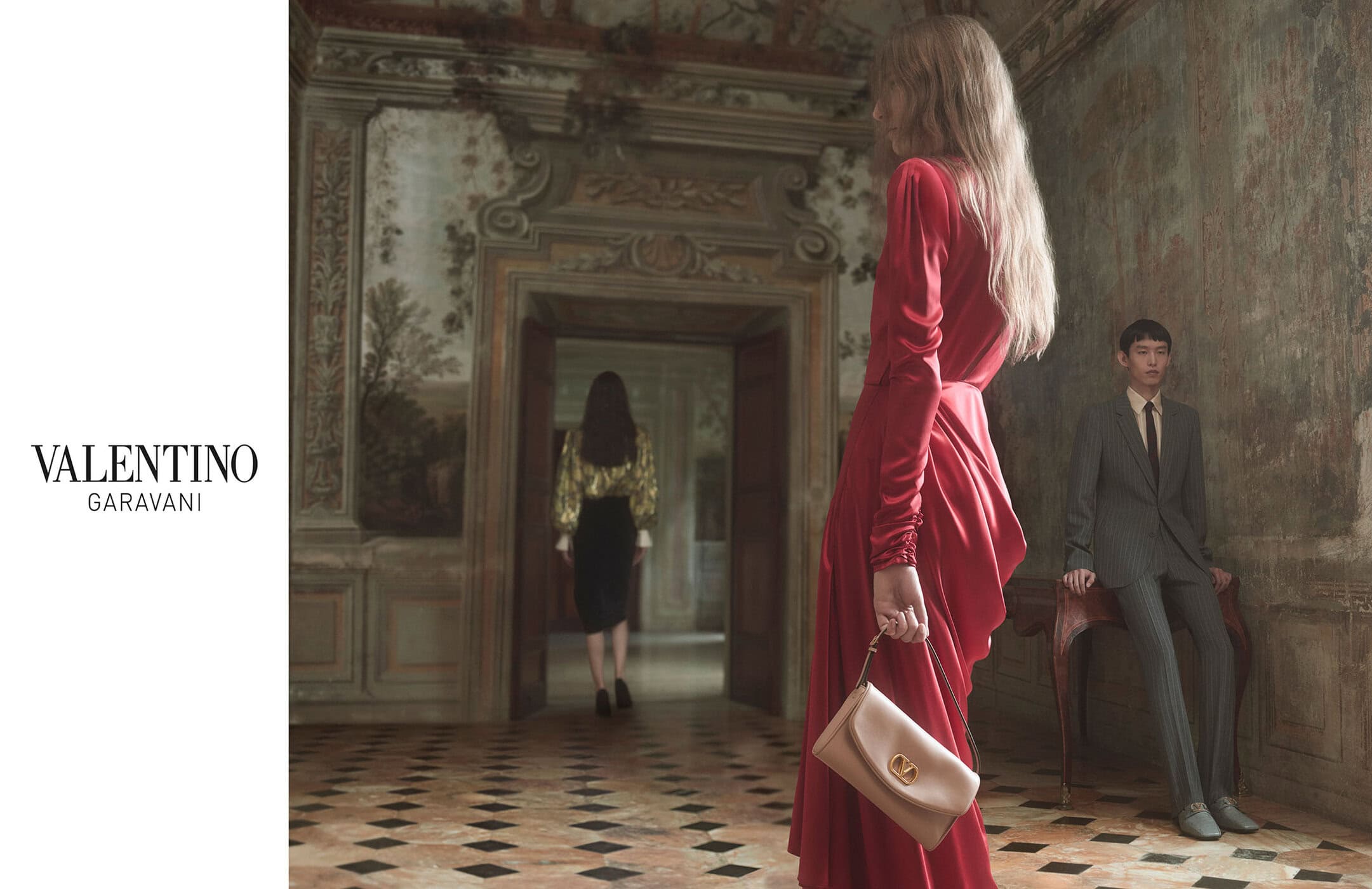 A woman in a red dress holds a purse in a grand interior. A man sits on a chair in the background, with another woman in a doorway.