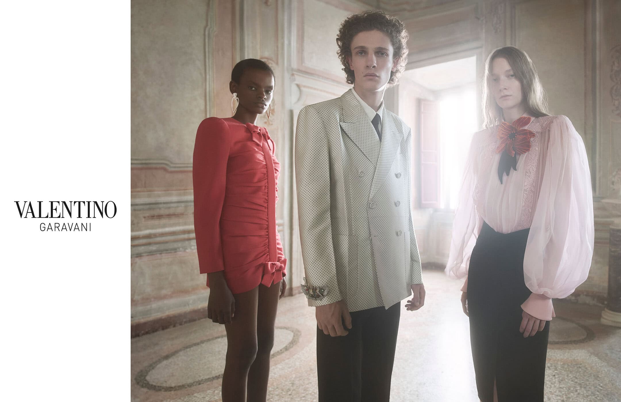 A fashion editorial shot of a group of three models, one male and two female, posing indoors, wearing formal dresses and suits.
