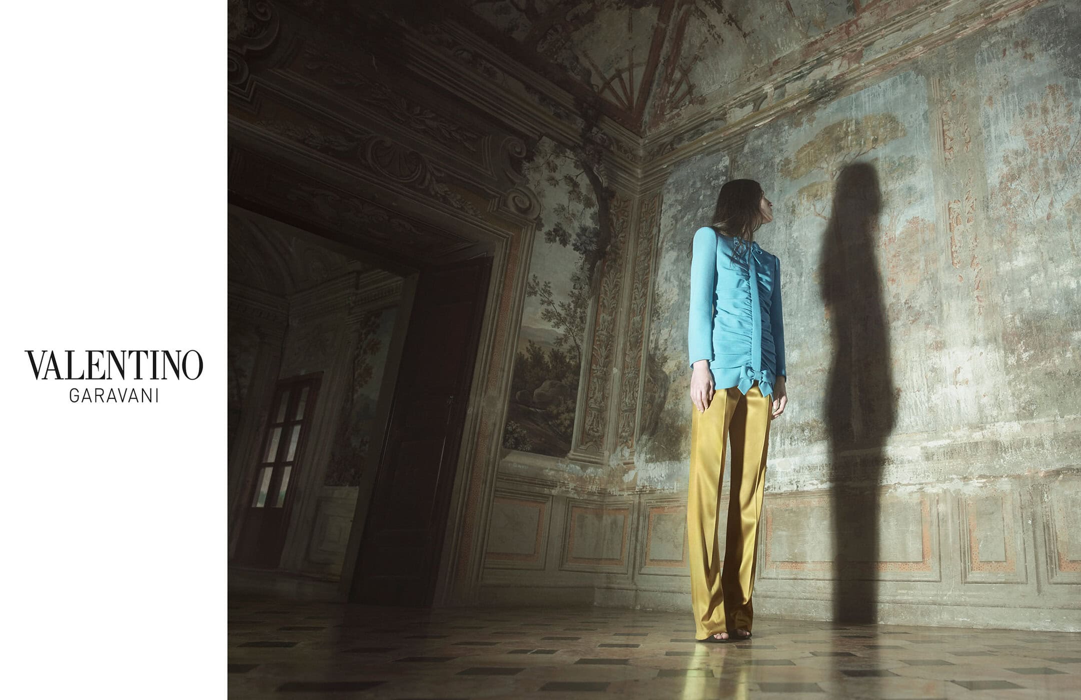 A person stands in an old, ornate room wearing a light blue shirt and yellow pants, the scene is lit with soft natural light.