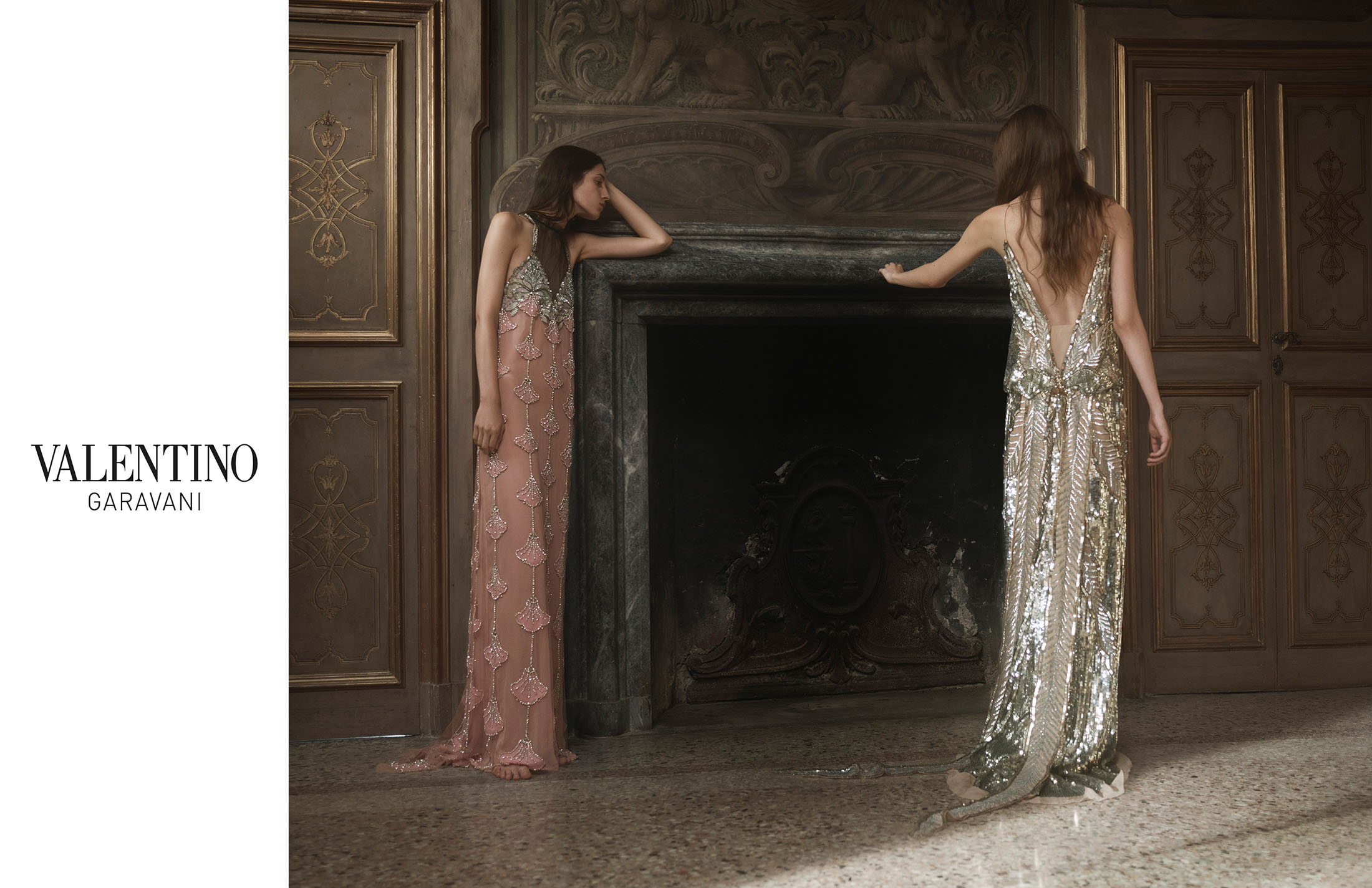 Two women in elegant gowns pose inside a grand hall. One woman faces away, showcasing the dress's back, in a Valentino fashion shot.
