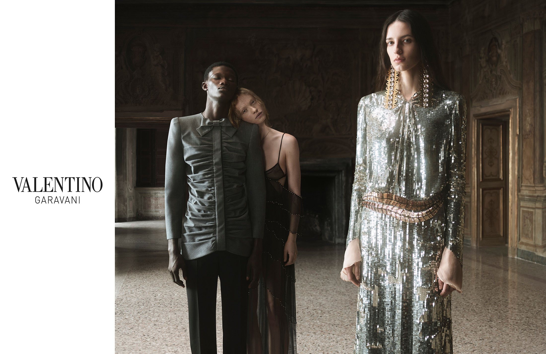 Fashion editorial shot of three fashion models, one in a silver sequin gown, inside a grand, ornate hall, promoting Valentino Garavani.