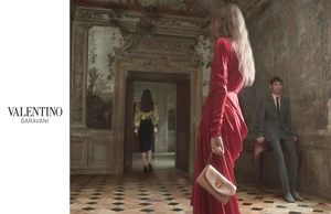 Model in a long red silk dress holding a tan handbag inside a classic room with frescoed walls and tiled floors, with Valentino logo on white side bar.