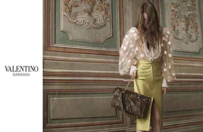 Model wearing sheer polka dot blouse and yellow skirt holding a brown leather handbag against an ornate classical wall