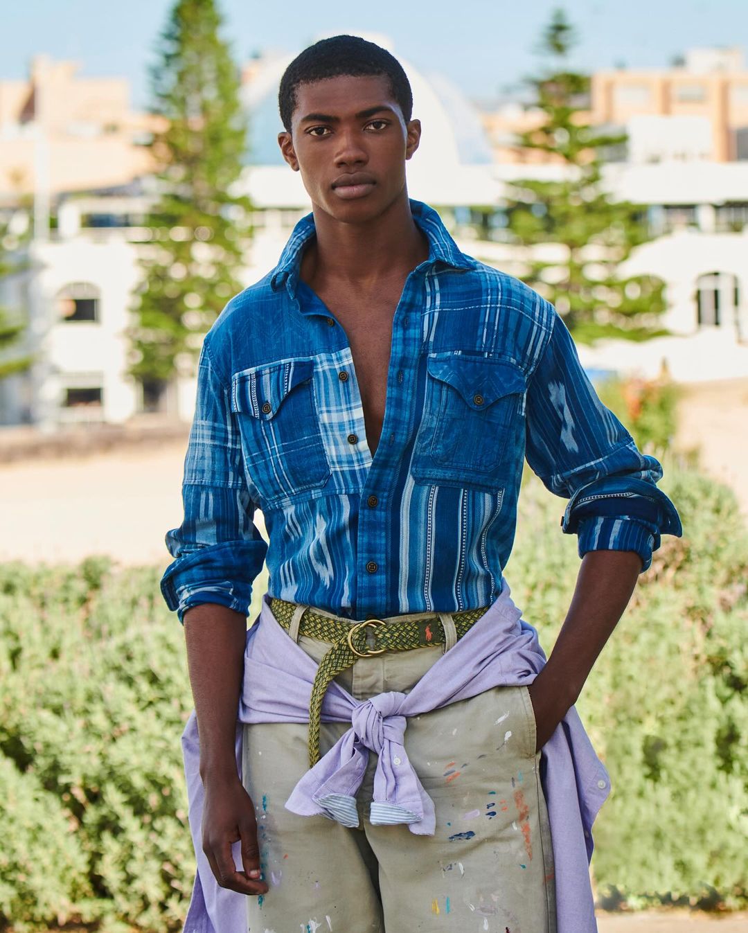 A young adult man with dark skin and dark hair is posing outdoors wearing a blue patterned shirt, khaki pants, and a braided belt.