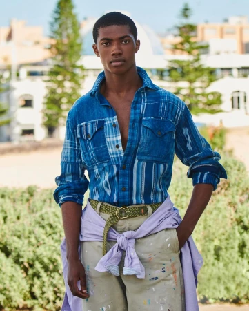 A young adult man with dark skin and dark hair is posing outdoors wearing a blue patterned shirt, khaki pants, and a braided belt.