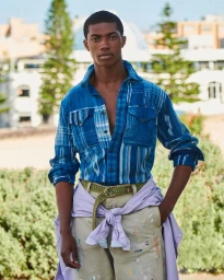 Black man wearing a blue plaid shirt and light trousers outdoors against a bright resort-style background for Esquire Summer 2024.