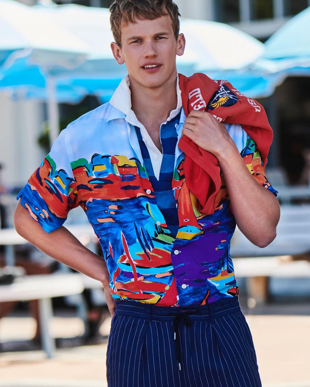 Handsome male model posing in a vibrant, patterned short-sleeve shirt and striped pants on a sunny day by the beach with blue umbrellas.
