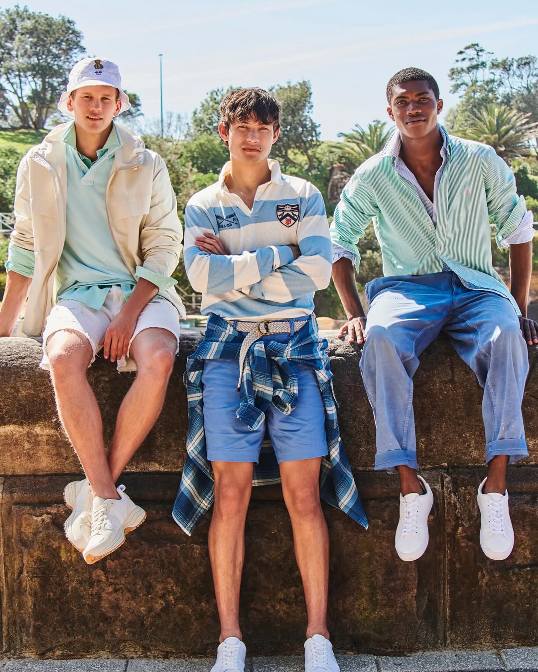 Three young men in casual summer fashion sit outside on a sunny day, showcasing classic and modern styles with a backdrop of greenery.
