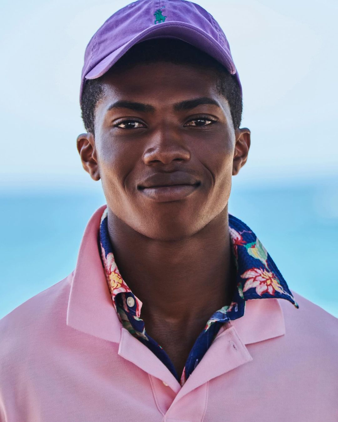 Close-up of a smiling African American male model wearing a purple baseball cap and pink short-sleeved shirt in front of a blurred blue backdrop.