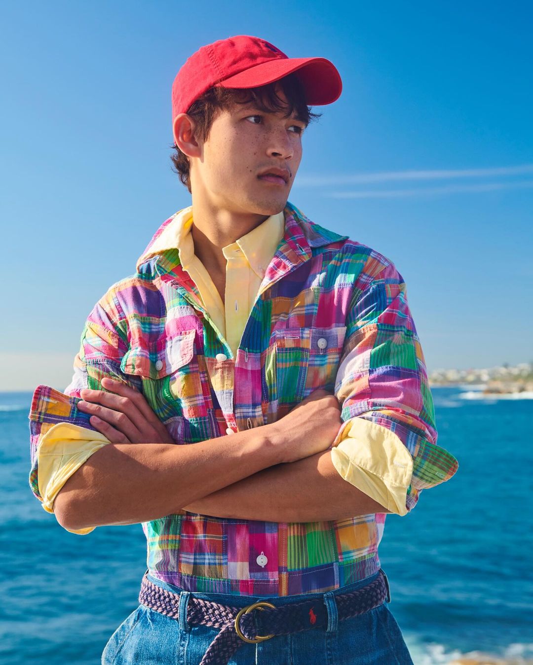A stylish young man in a colorful plaid shirt, red cap, and blue jeans standing outside with a bright blue sky and ocean backdrop.