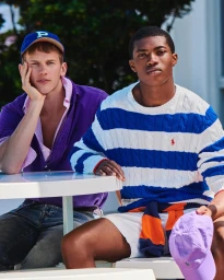 Two male models seated at a white table outdoors wearing casual summer apparel including a blue cap and striped sweater for Esquire.