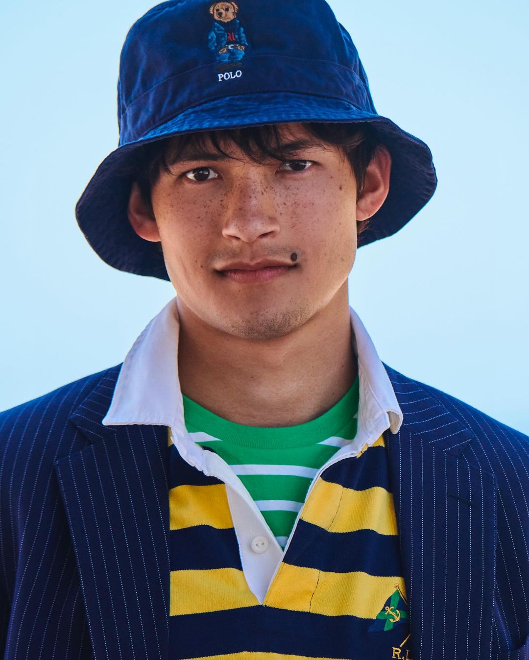Close-up of an adult male wearing a blue bucket hat with a teddy bear logo, pinstripe blazer, and striped polo shirt in an outdoor setting.
