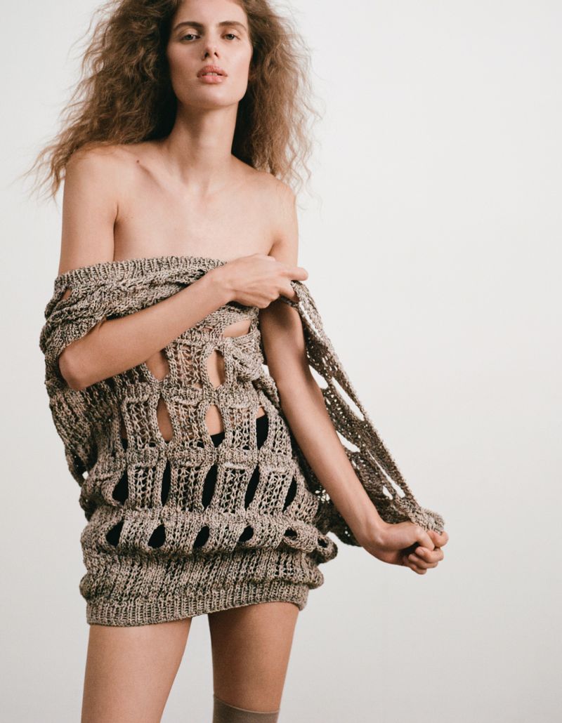 Woman in a knitted open-weave mini dress poses in front of a white background in a fashion shot emphasizing textures and neutral tones.