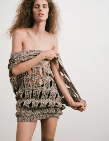 Woman in a knitted open-weave mini dress poses in front of a white background in a fashion shot emphasizing textures and neutral tones.