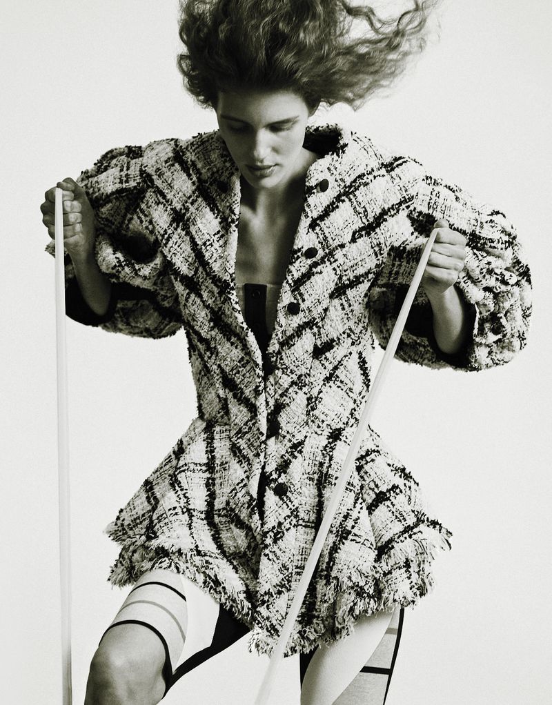 Monochrome studio shot of a woman with voluminous hair wearing a textured blazer and striped tights while holding two white rods.