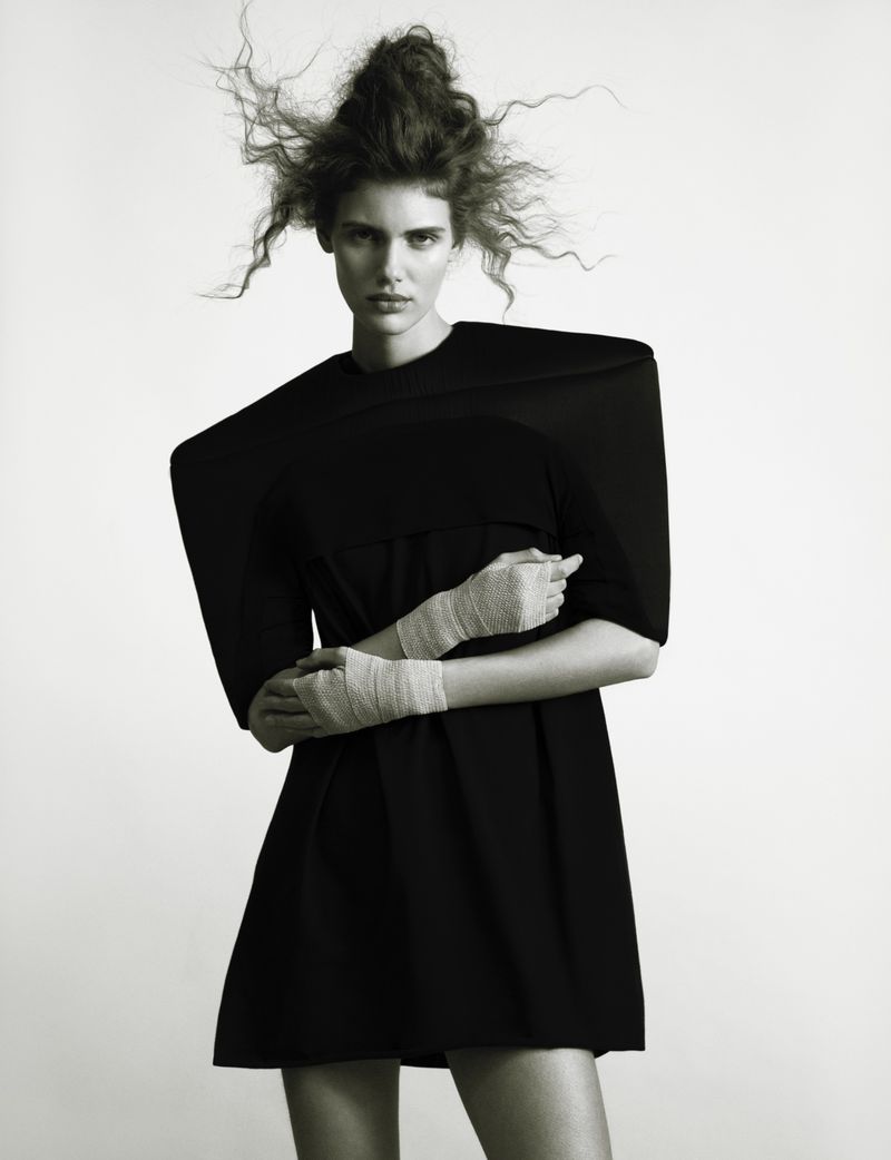 Studio shot of a model wearing a black dress with exaggerated shoulders, arms crossed with wrapped bandages in a high-key setting.