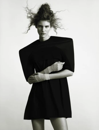 Studio shot of a model wearing a black dress with exaggerated shoulders, arms crossed with wrapped bandages in a high-key setting.