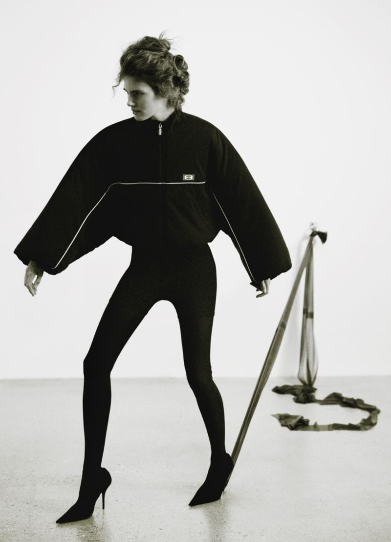 A woman in black athletic-style clothing poses in front of a white background with a ballet barre and fabric in a minimalist studio setting.