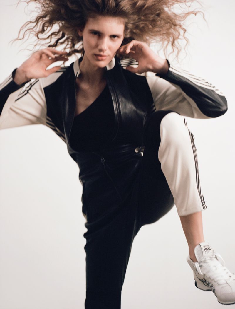 Fashion shot of a young woman with curly hair wearing an athletic-style outfit with black and white details and white sneakers, studio lit.
