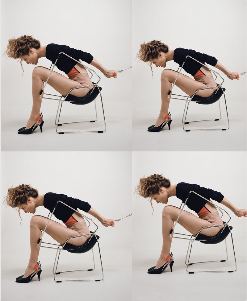 Woman in black top and heels sits backward in a black chair with a chrome frame against a light background in a studio setting.