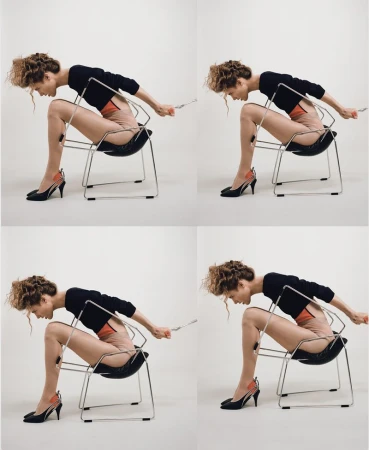 Woman in black top and heels sits backward in a black chair with a chrome frame against a light background in a studio setting.