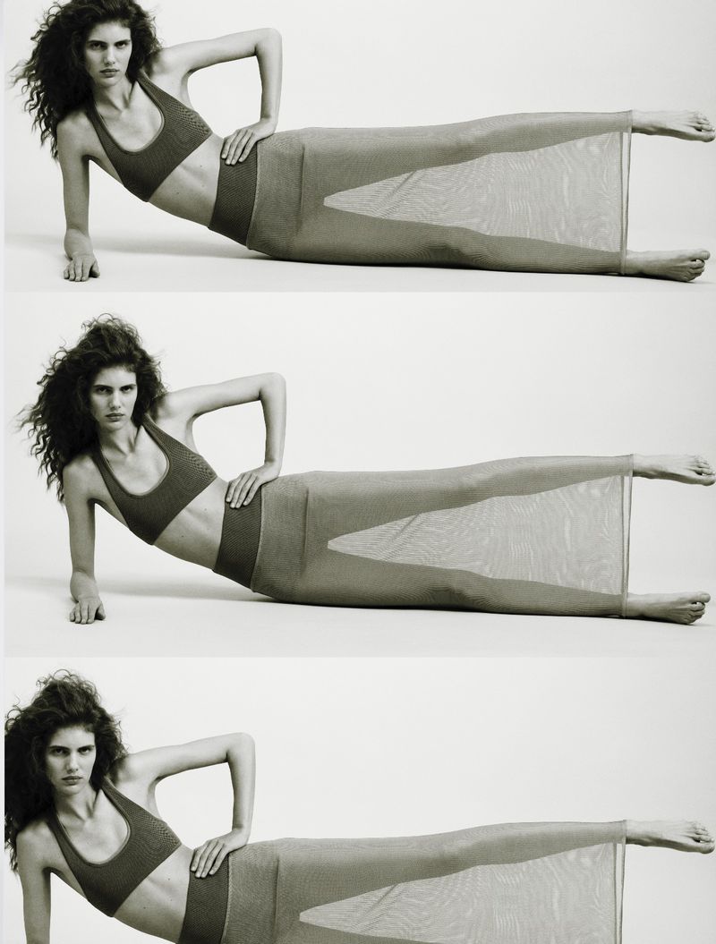 Studio shot of a woman with curly dark hair, lying on her side wearing a dark sports bra and skirt, with a neutral white background.
