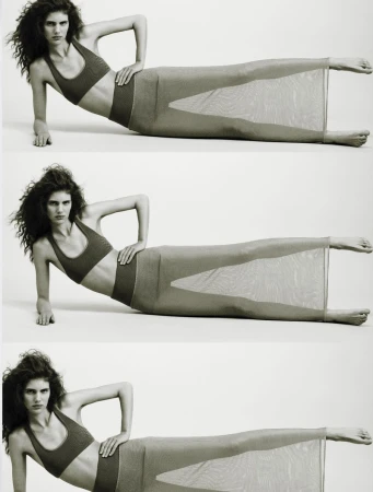 Studio shot of a woman with curly dark hair, lying on her side wearing a dark sports bra and skirt, with a neutral white background.