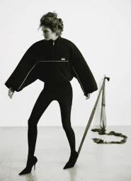 Model wearing a black zip jacket and black tights posing against a white studio background with a hanging ribbon prop