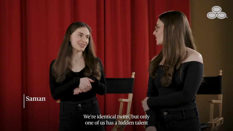 Two adult women pose in a medium indoor shot with a red curtain backdrop. Both wear dark clothing and have long brown hair in studio.
