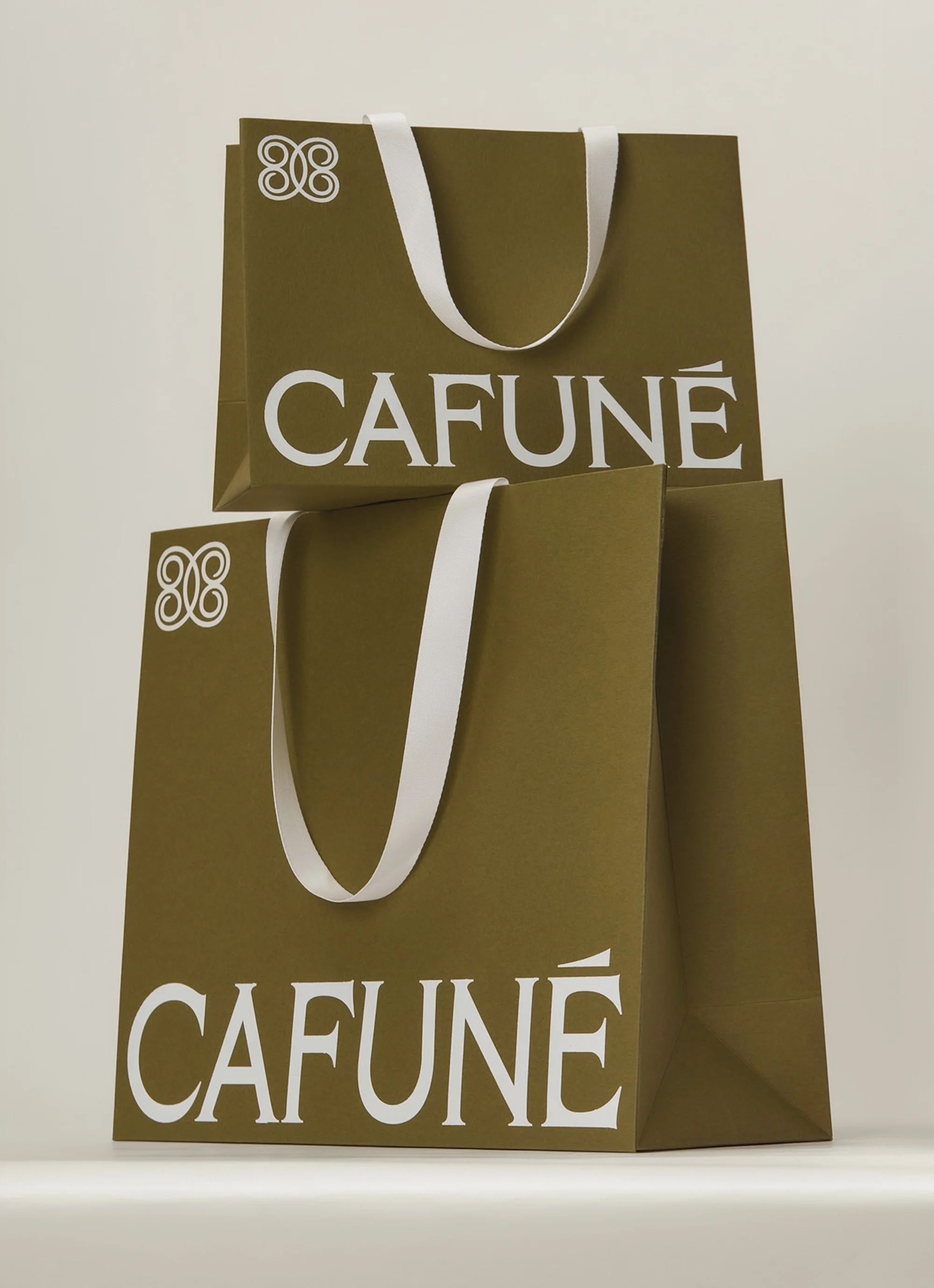 Two tan paper shopping bags from CAFUNE with white handles, stacked against a neutral background in a studio-lit, eye-level shot.