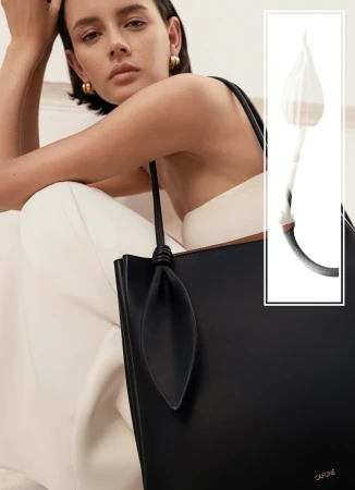 Model with short dark hair wearing gold earrings and holding a black leather tote bag against a neutral background.